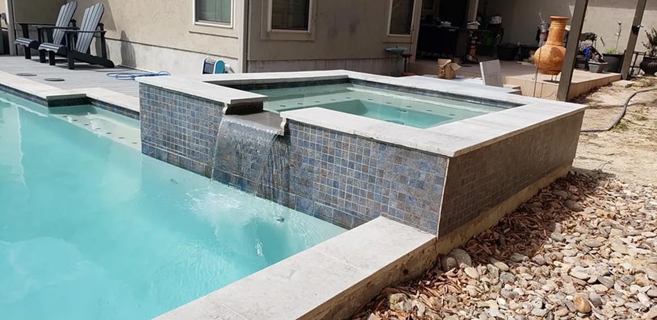 Hot tub seen with a waterfall pouring into the main pool, house can be seen in background